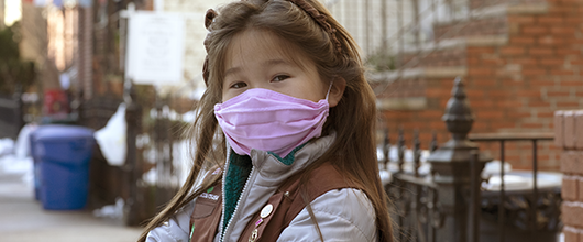 A Girl Scout wears a brownie vest and a light purple, surgical face mask.