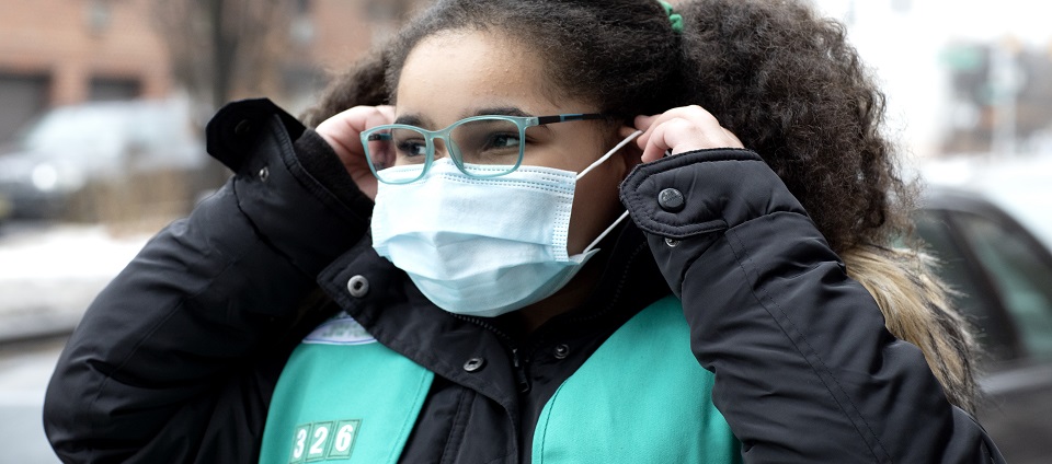 A Girl Scout in a green vest pulls a surgical mask on over her face.