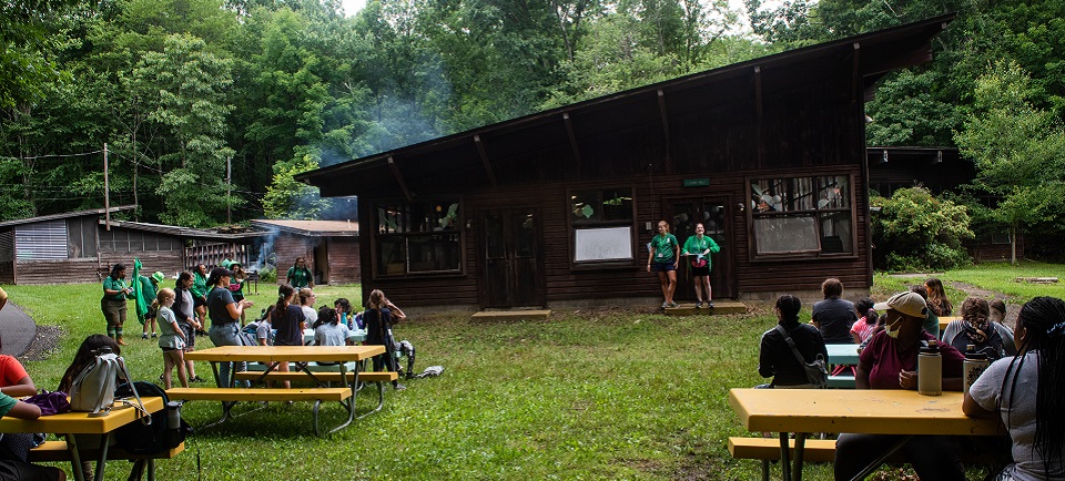 Girl Scouts gathered at picnic tables at Camp Kaufmann. Two counselors makes accounements while the smoke from grilling burgers rises in the background.