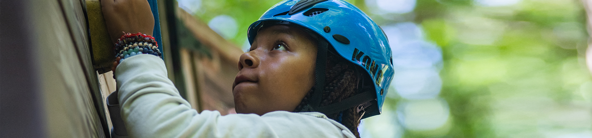  A younger Girl Scout with a helmet on at the high ropes course at camp, surrounded by other Girl Scouts and counselors 