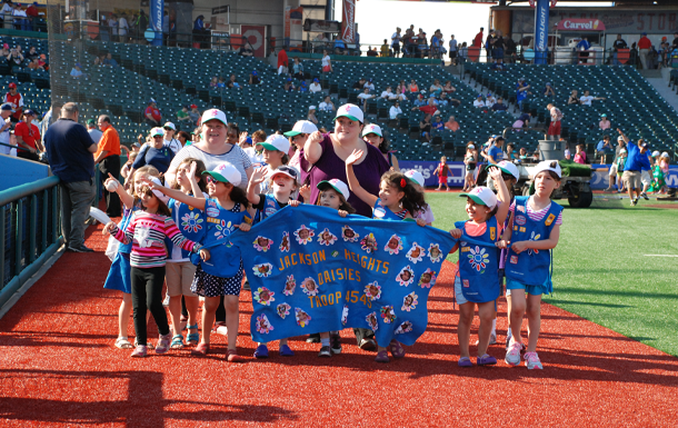 A troop of daisy Girl Scouts and their two troop leaders walking with a banner around the field of a baseball stadium