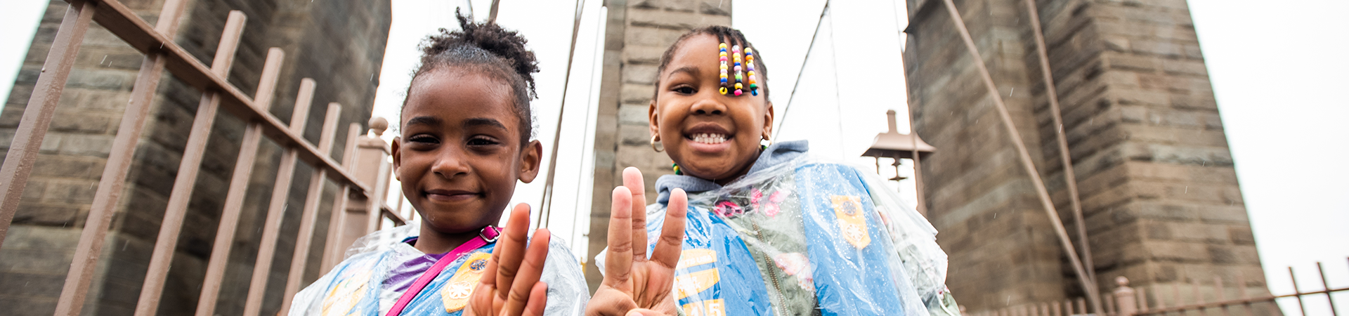  two daisy girl scouts on the brooklyn bridge, smiling holding up the girl scout sign with their hands 