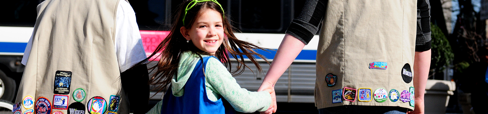  adult volunteer wearing vest with a girl scout junior (right, in baseball cap) and daisy (left) outside at a park smiling and looking at one another 