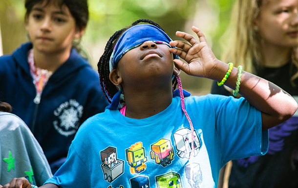 Girl Scout at camp looking straight up into the air with a blindfold over their eyes, reaching up with their hand to pull it off.